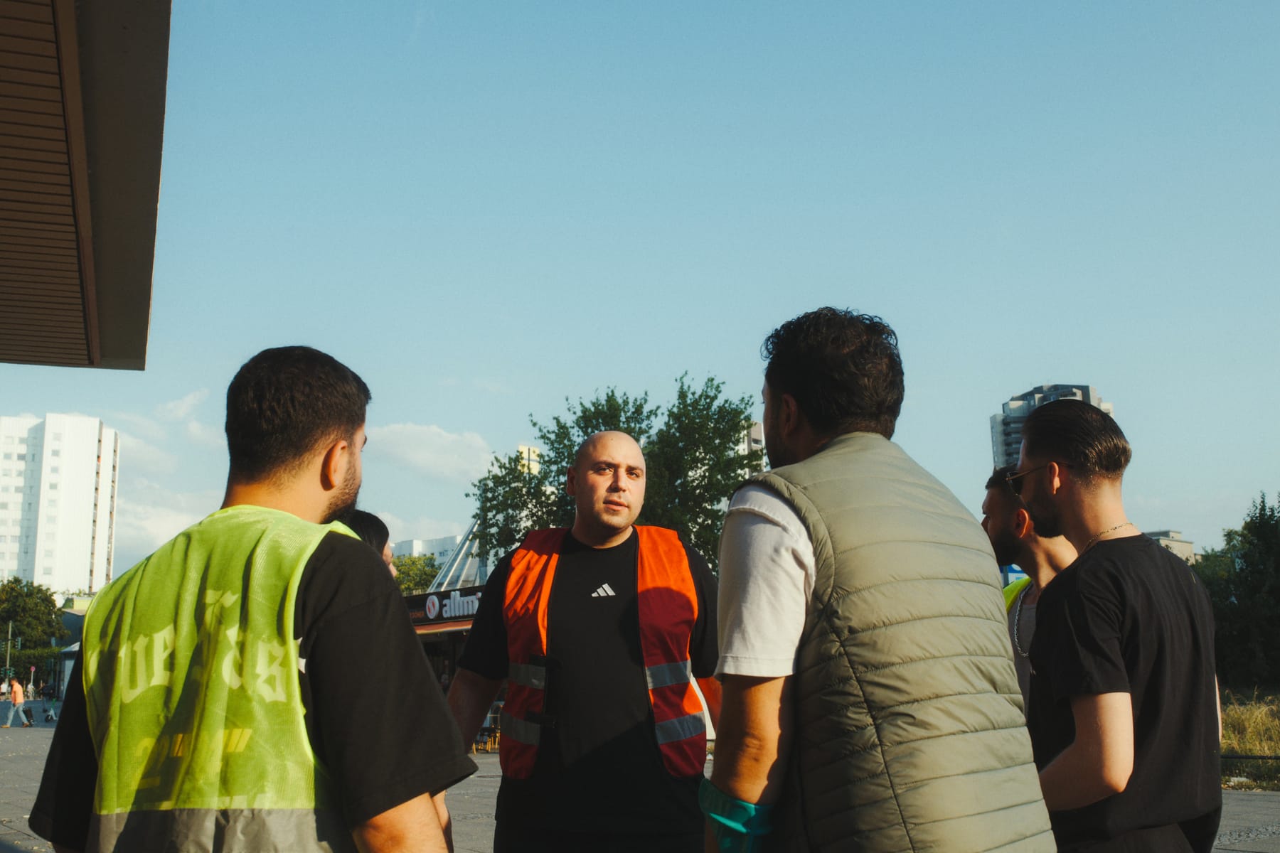 Yurtsever Özdemir in Warnweste im Gespräch mit jungen Männern, Hochhäuser der Gropiusstadt im Hintergrund